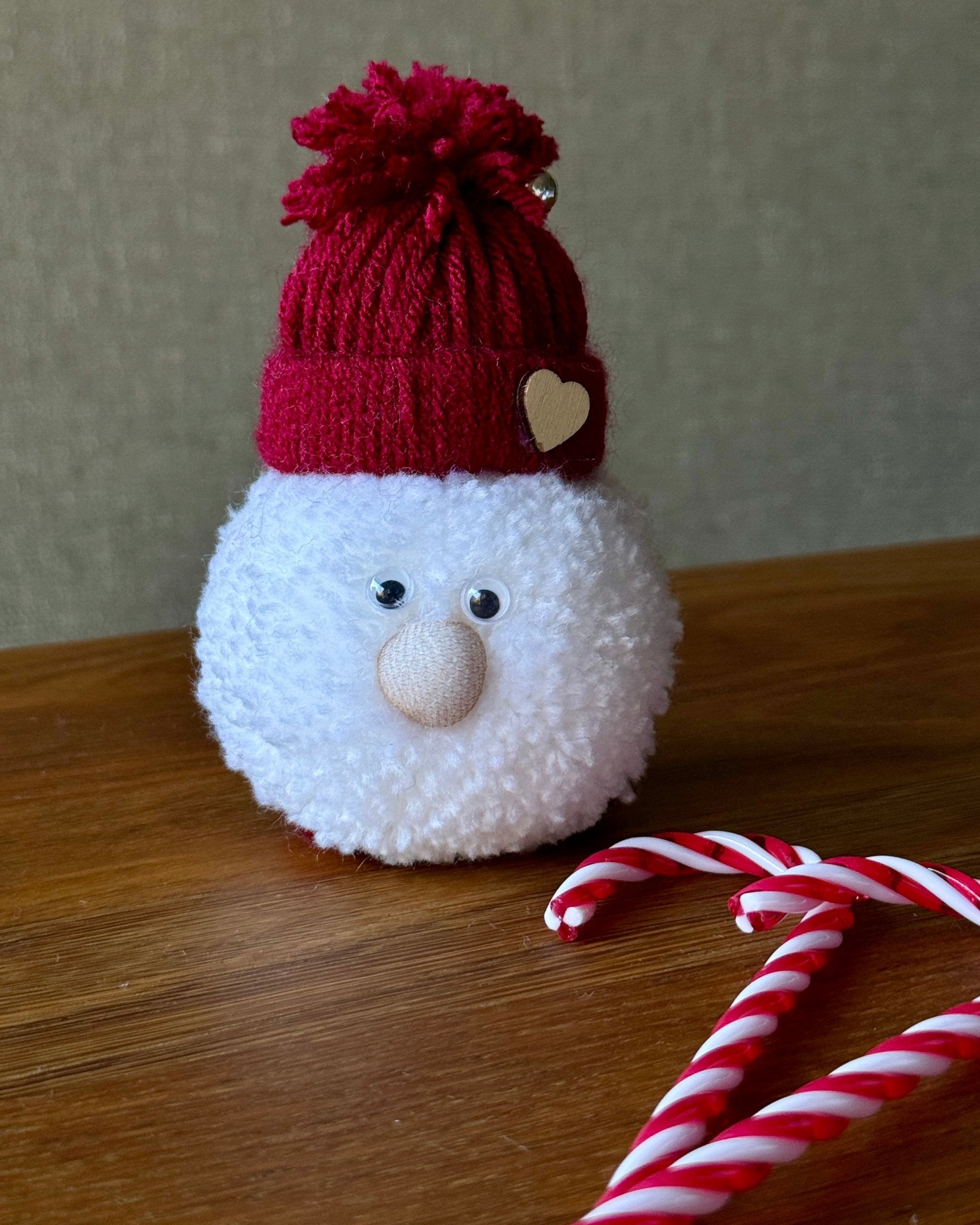 Mehka okrasna snežna figura s kapo v bordo rdeči barvi in cofom, postavljena na leseno površino.