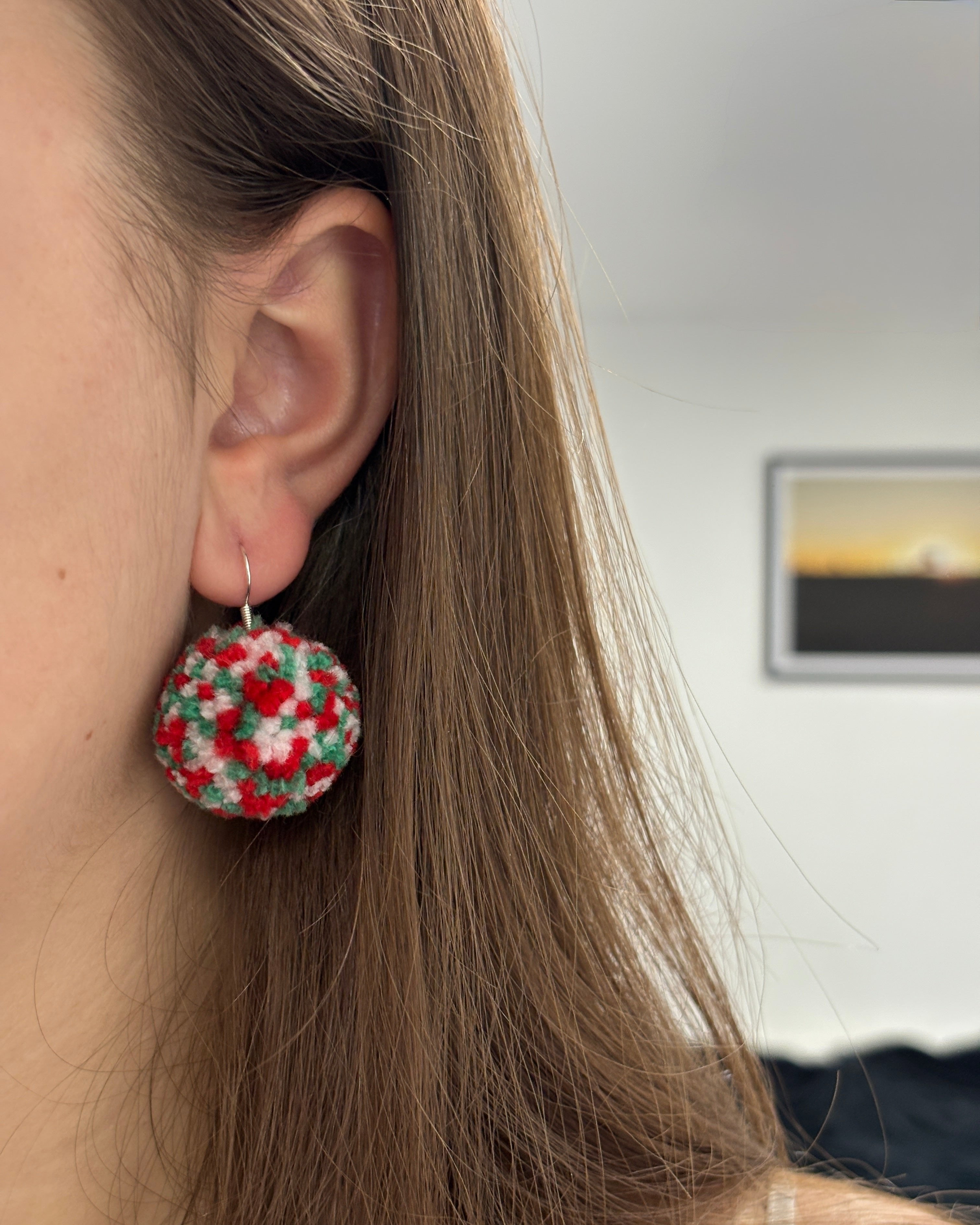 Belo-rdeče-zeleni pom-pom viseči uhani v ušesu ženske kot modni dodatk, v ozadju slika na steni.