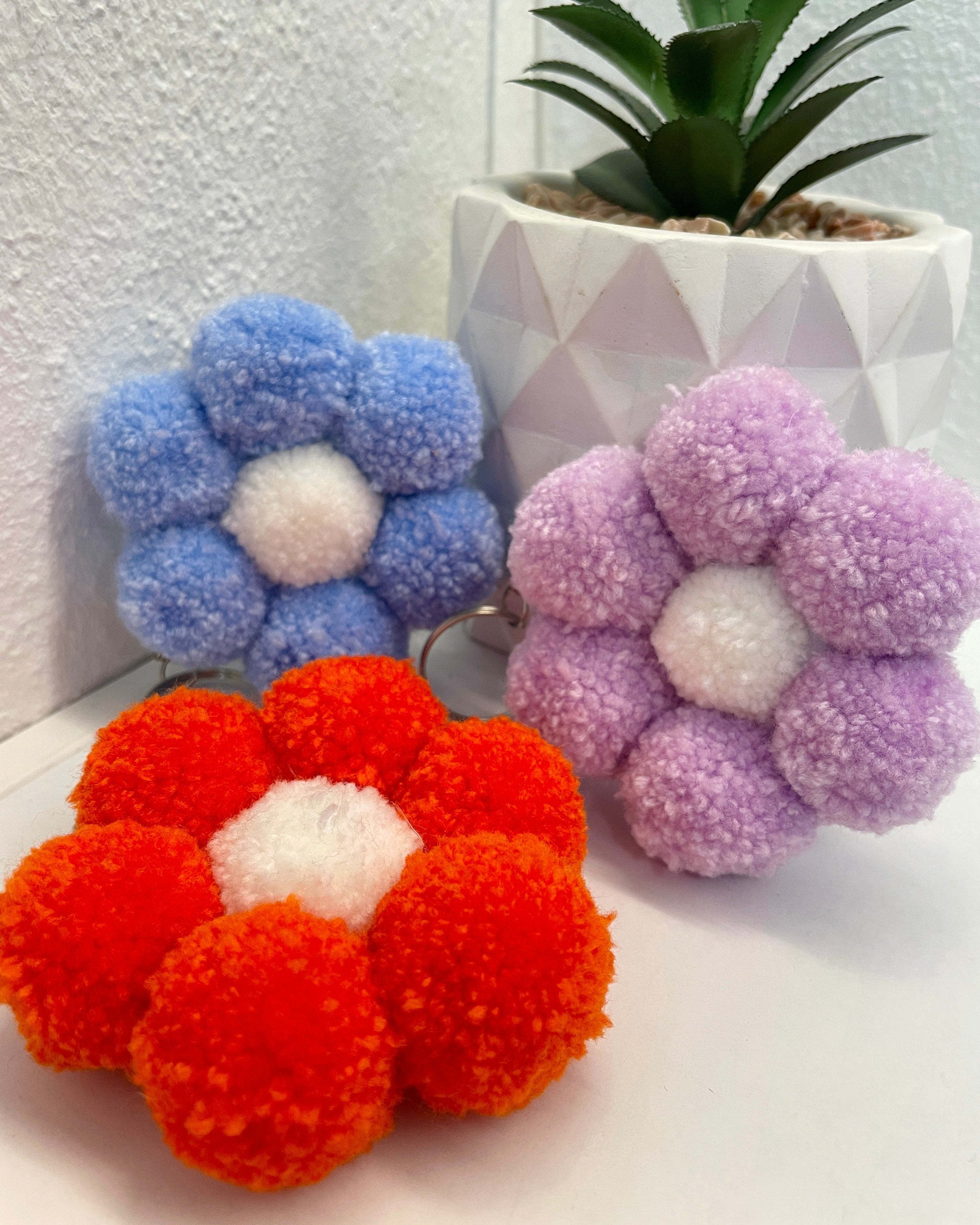 Pom pom obesek v obliki rože, na beli površini, z rožo v ozadju
