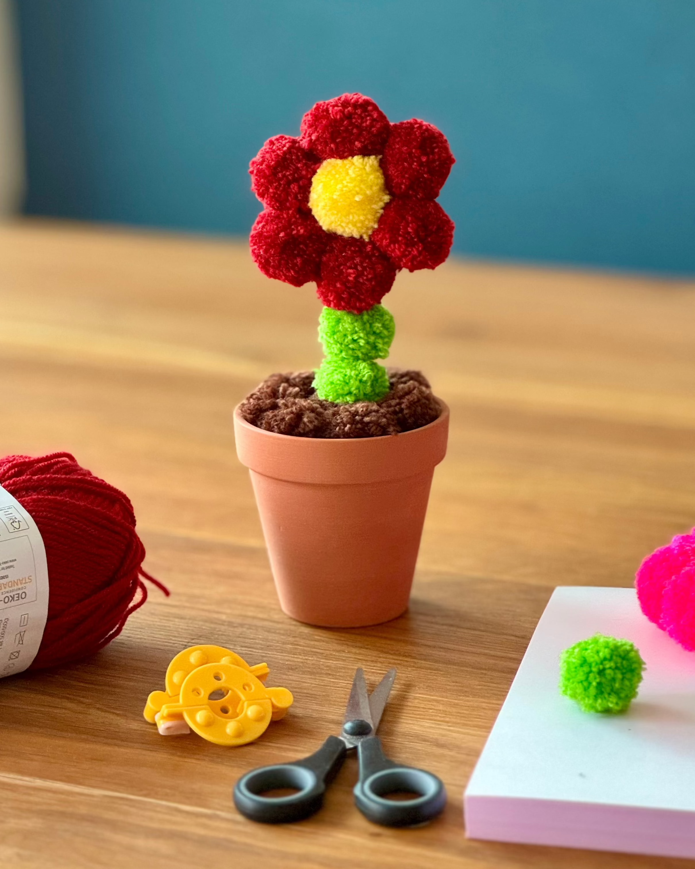 Dekoracija na leseni mizi, roža iz cofov pom pom v glinenem lončku, preja volne, škarje, pripomočki za izdelavo cofov