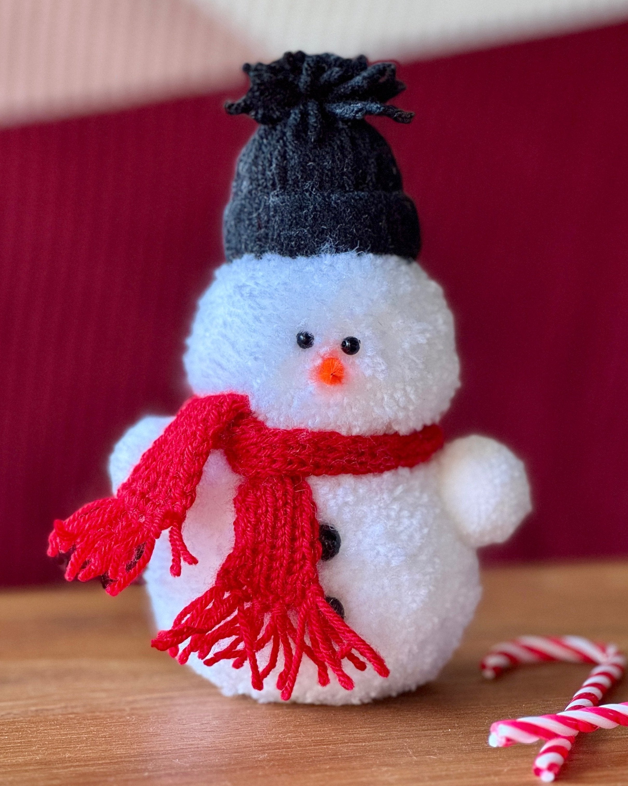 Snežak iz cofa pom pom, s črnim šalom in rdečo kapo, postavljen na mizo, črni gumbi, sladkorna palčka, božično ozadje.