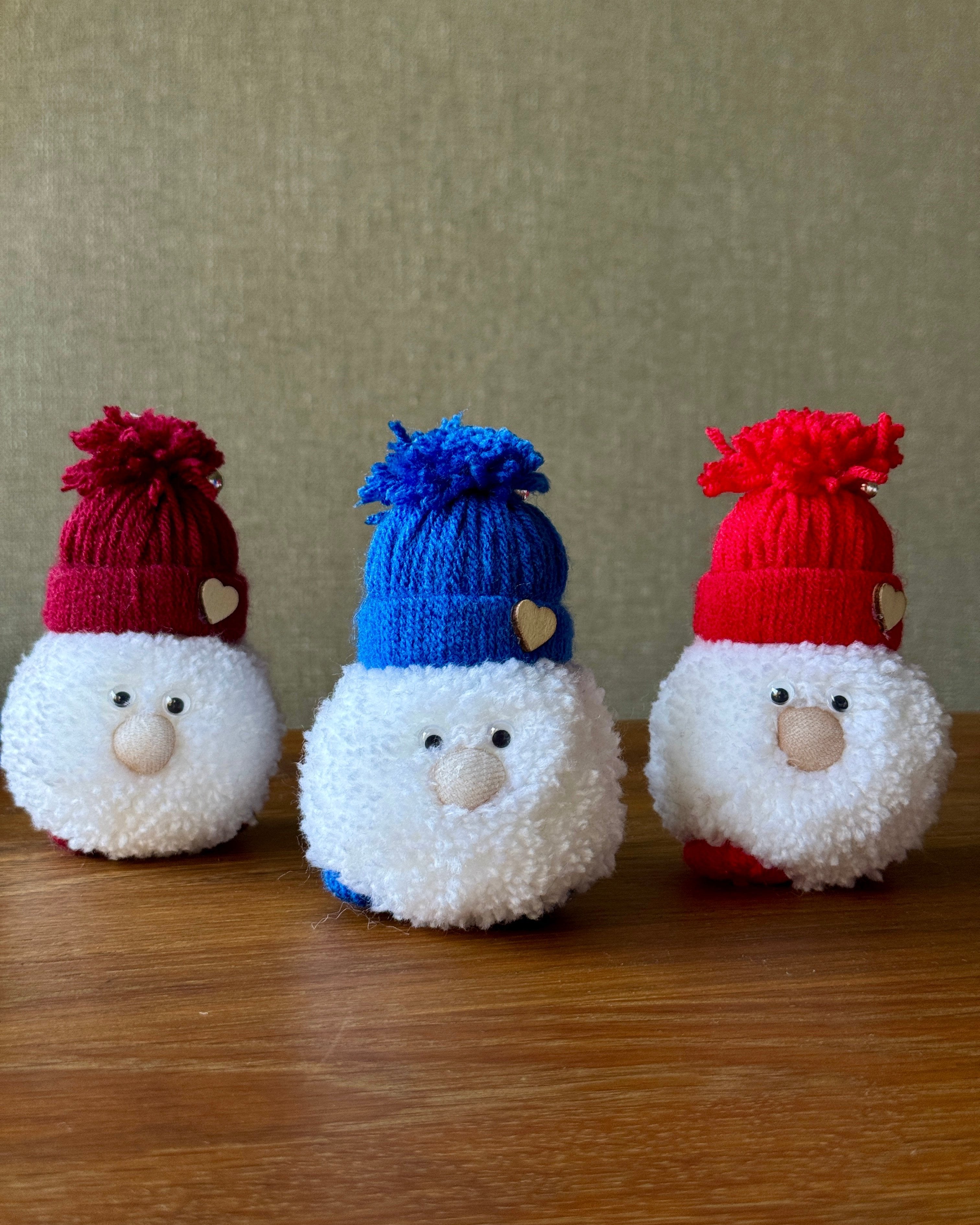 Mehka okrasna snežna figura s kapo v različni barvi in cofom, postavljena na leseno površino.