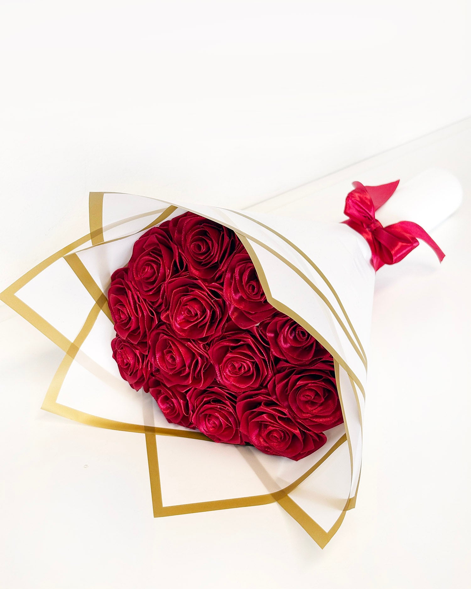 Šopek satenastih vrtnic v bordo rdeči barvi, zavite v bel korejski papir z zlatim robom, z bordo rdečo pentljo
